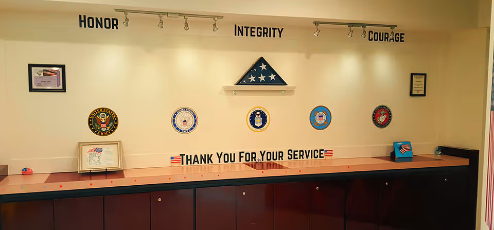 A wall display in a retirement facility featuring the words 'HONOR', 'INTEGRITY', and 'COURAGE' above military emblems and a folded American flag. Below the emblems is a sign that reads 'THANK YOU FOR YOUR SERVICE' with small American flag icons on either side. The display is set above a wooden cabinet with a light-colored wall background.