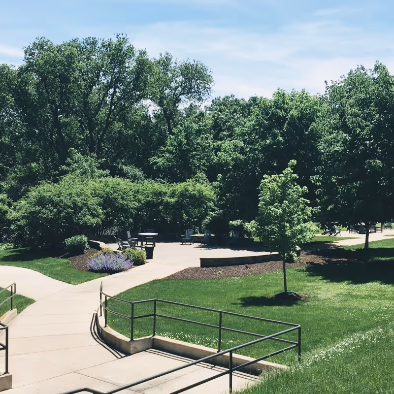 Outdoor area with a paved walkway and seating area surrounded by green grass, trees, and bushes under a partly cloudy sky.