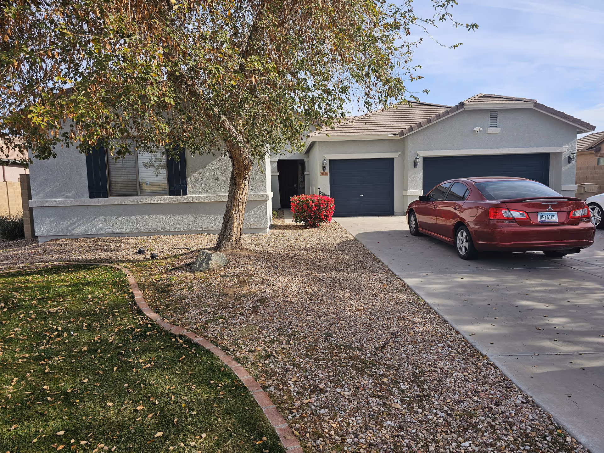 Front exterior of a single-story stucco house with a two-car garage, a red sedan in the driveway, gravel landscaping, and a tree by the entrance.