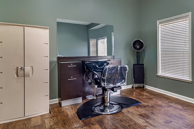 A small salon area with a black salon chair covered in plastic in front of a dark wooden counter with a large mirror. To the left is a beige storage cabinet, and to the right is a window with closed blinds and a black hair dryer on a stand. The room has green walls and wood flooring.