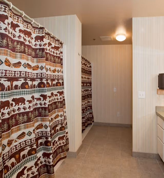 Interior view of a bathroom area with two shower stalls separated by a wall. Each shower has a curtain featuring a rustic pattern with bears, moose, trees, and canoes in brown, green, and orange colors. The floor is tiled, and the walls have light-colored vertical striped wallpaper. A ceiling light is visible above.