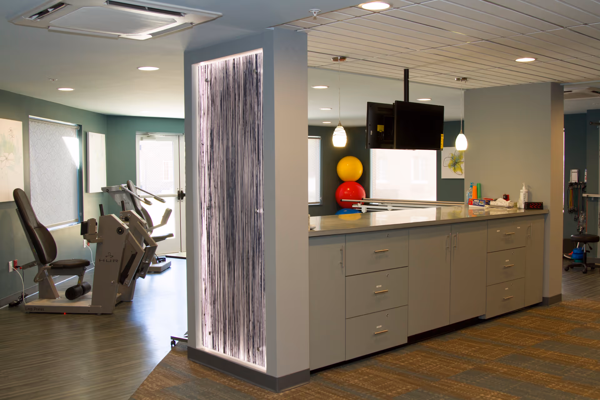 Interior view of a fitness or rehabilitation room in a senior living facility, featuring exercise equipment such as a leg press machine, a countertop with cabinets, a mounted TV, and colorful exercise balls in the background. The room has a modern design with wood flooring and soft lighting.
