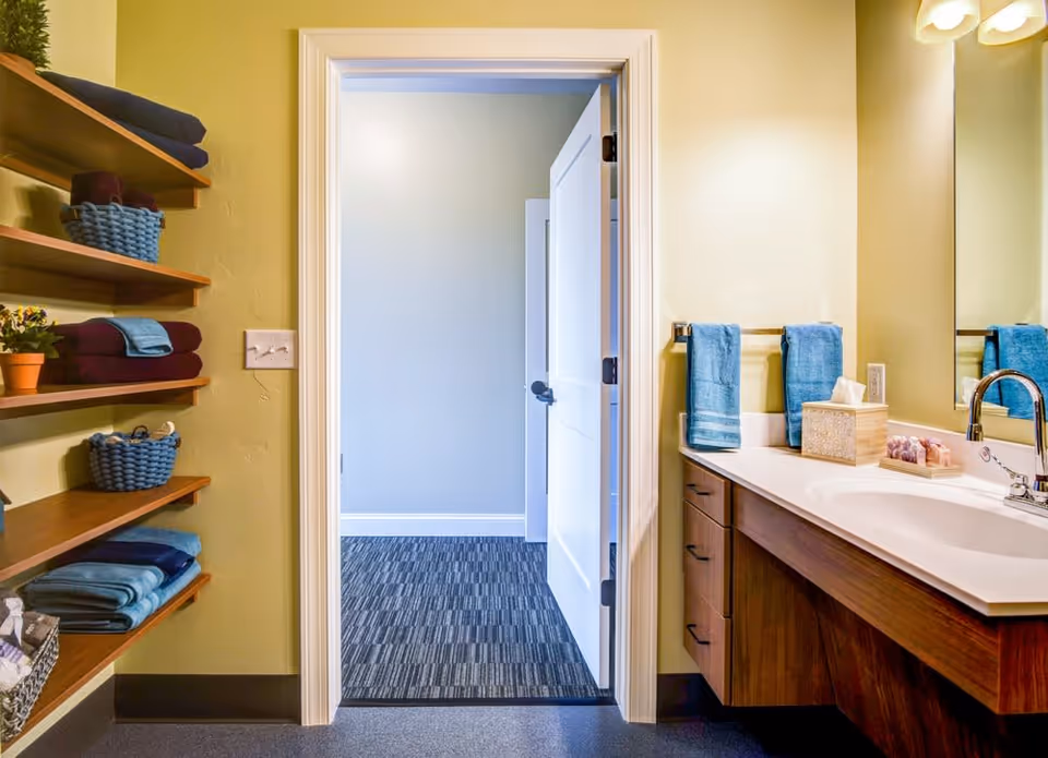 A bathroom interior with a wooden vanity and white countertop featuring a sink and faucet. Two blue hand towels hang on a towel rack above the counter, which also holds a tissue box and soap. To the left, wooden shelves hold neatly folded towels and small baskets. The bathroom door is open, revealing a carpeted room beyond with light-colored walls.