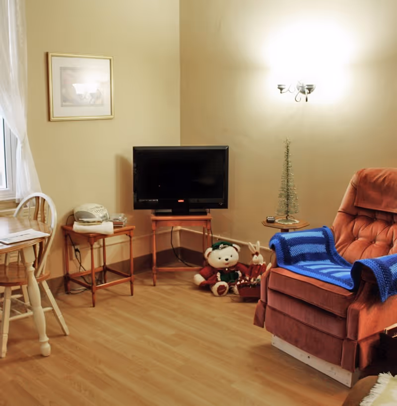 A cozy living room corner with a brown recliner chair draped with a blue crocheted blanket, a small wooden table holding a flat-screen TV, a teddy bear and a small stuffed rabbit on the floor, a small decorative Christmas tree on a side table, and a wooden chair and table near a window with sheer white curtains.
