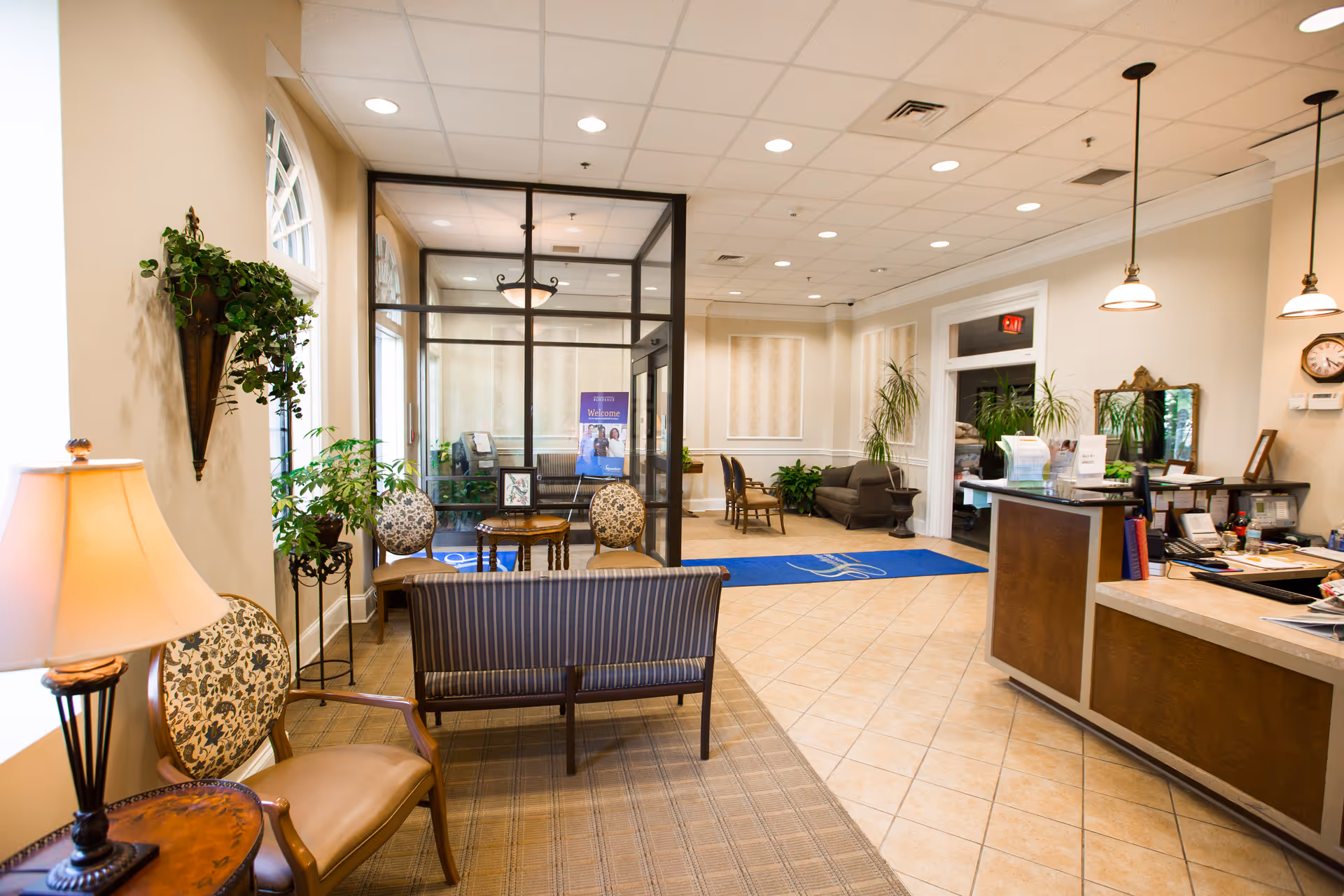 A bright and welcoming reception area of a senior living facility with a front desk on the right, seating area with chairs and a small table on the left, potted plants, and a glass-enclosed office or room in the background. The floor is tiled, and the walls are painted in light colors with decorative moldings. There are hanging pendant lights above the reception desk and a blue welcome mat near the entrance.