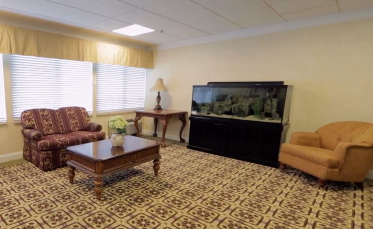 A cozy living room with patterned carpet, a striped upholstered loveseat, a wooden coffee table with a flower vase, a side table with a lamp, a large fish tank, and a cushioned armchair.