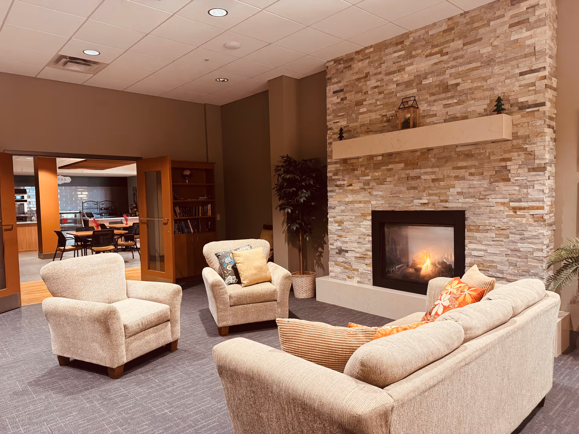 A cozy living room area in an assisted living facility featuring a beige sofa and two matching armchairs with decorative pillows arranged around a modern stone fireplace with a fire burning. In the background, there is a doorway leading to a dining area with tables and chairs.