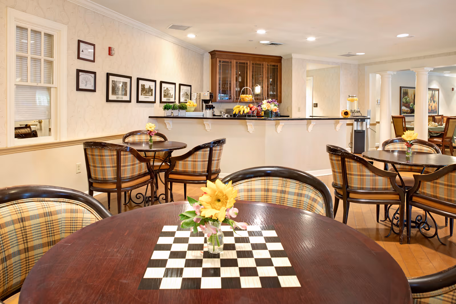 A cozy dining area in a senior living facility with round wooden tables and plaid cushioned chairs. Each table has a small vase with a yellow flower. In the background, there is a counter with fruit baskets and a coffee machine, along with framed pictures on the wall.