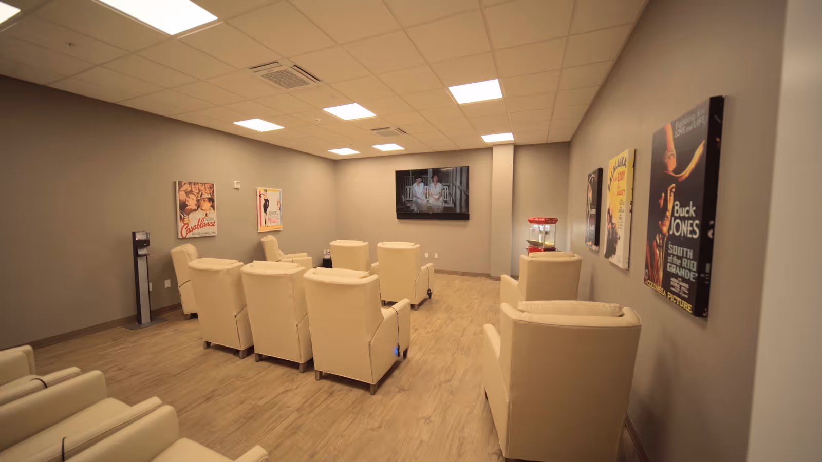 Small screening room with rows of beige recliner chairs facing a wall-mounted TV, movie posters on the walls, and a popcorn machine.