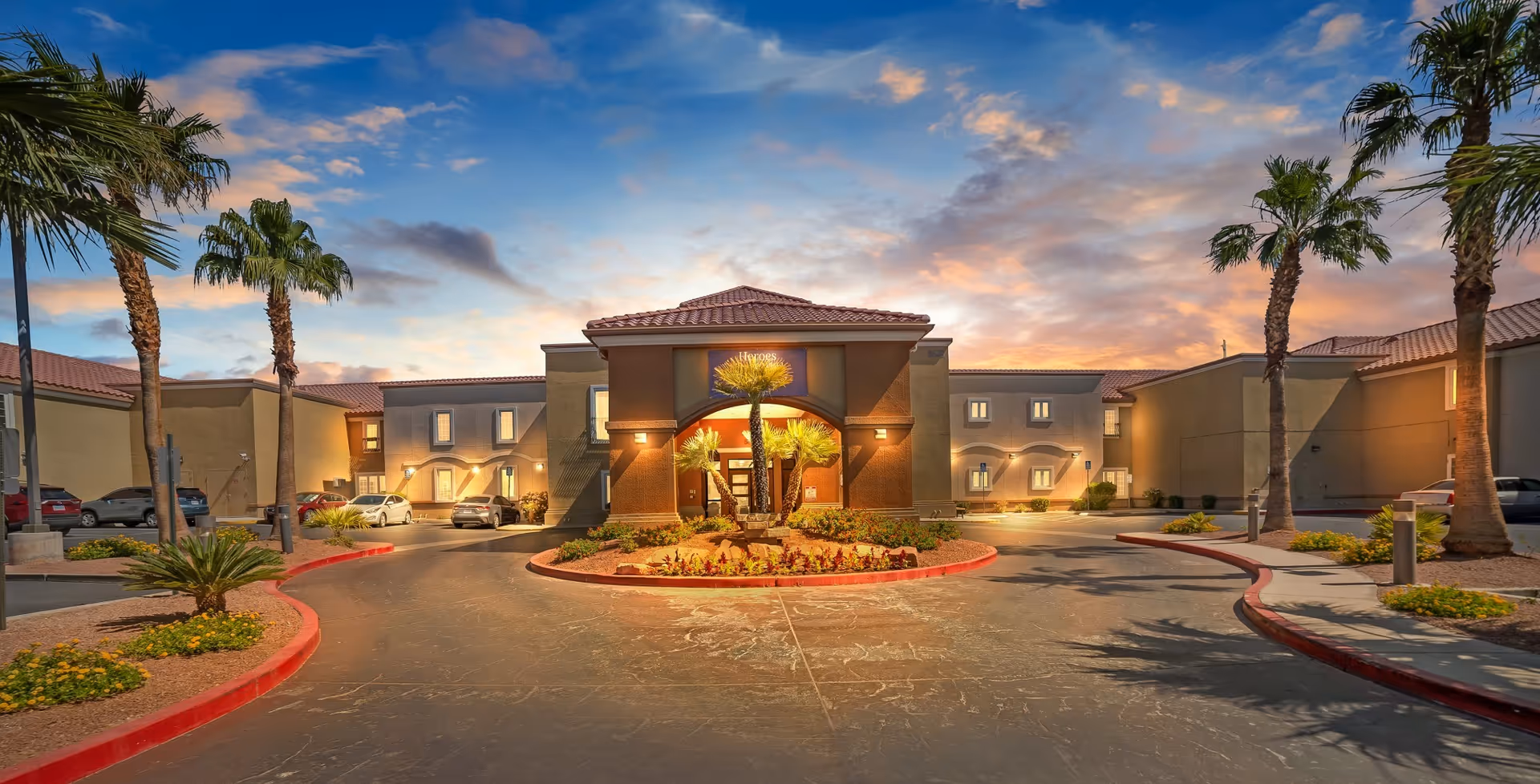 Exterior view of Acacia Springs senior living facility at sunset with a circular driveway, palm trees, and landscaped flower beds in front of the building entrance.