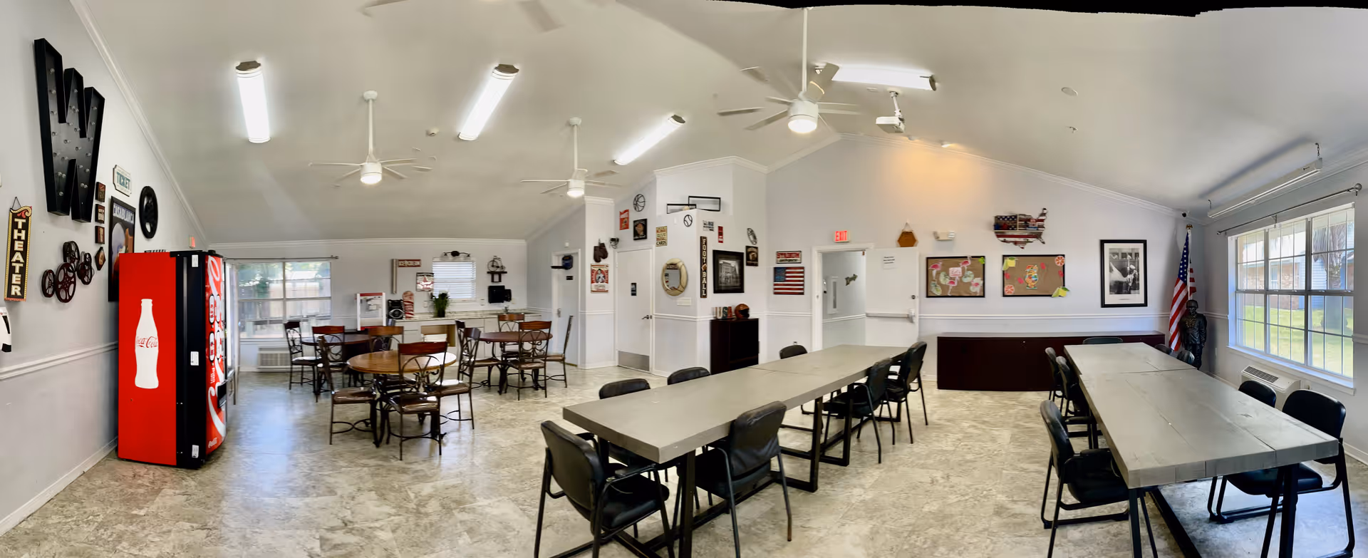 A spacious common room with multiple tables and chairs arranged for group seating. On the left side, there is a red Coca-Cola vending machine next to a wall decorated with various signs and clocks. The room has large windows letting in natural light, white walls, and a light-colored floor. Ceiling fans and fluorescent lights are mounted on the ceiling. An American flag is displayed near the windows on the right side.