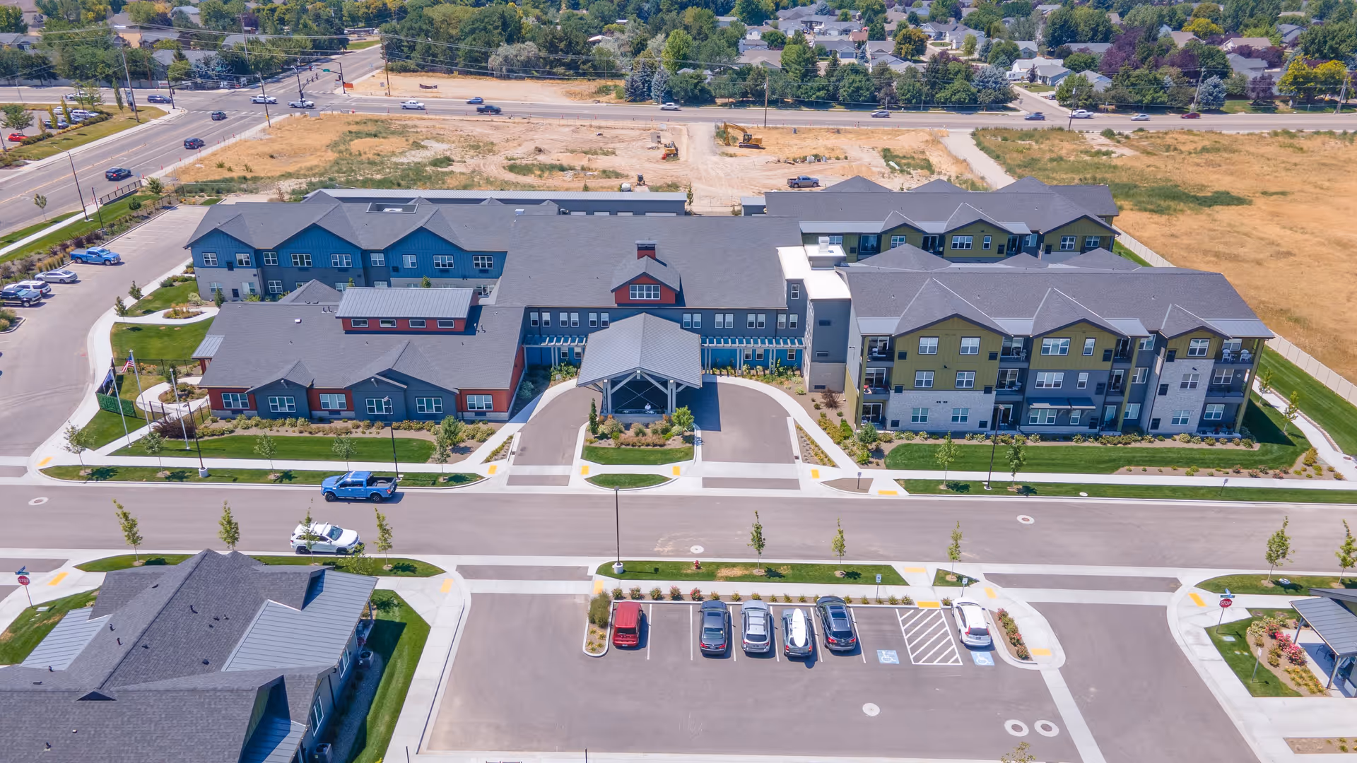 Aerial view of The Pointe at Meridian senior living facility showing a large multi-wing building with a covered entrance, surrounded by parking lots, roads, and landscaped green areas. Residential neighborhoods and undeveloped land are visible in the background.