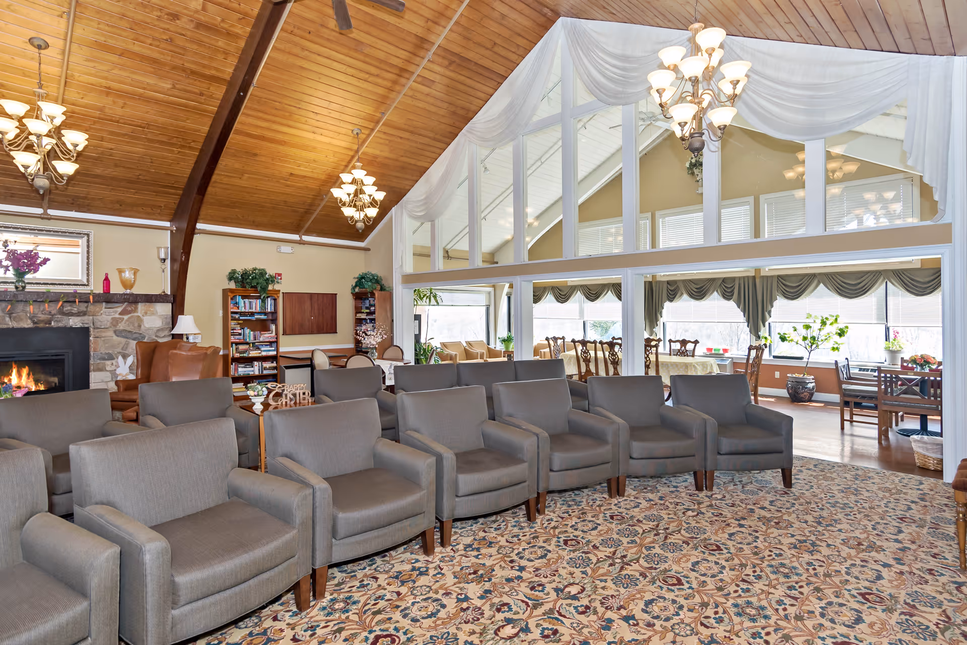 A spacious living room area with a high wooden ceiling and multiple chandeliers. There are two rows of gray armchairs arranged on a patterned carpet. In the background, there is a stone fireplace with a fire burning, a bookshelf, and a seating area with brown leather chairs. Large windows with white curtains separate this room from a sunlit dining area with tables, chairs, and plants.
