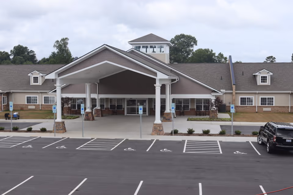 Front exterior view of Grace Village Assisted Living and Memory Care building with a covered entrance supported by white columns, several handicap parking spaces in front, and a black SUV parked on the right side.