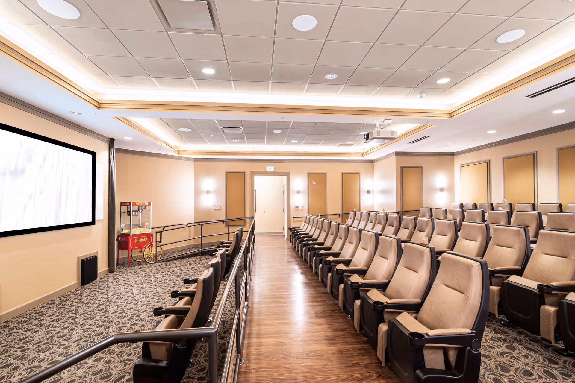 Interior view of a small theater room with multiple rows of beige cushioned seats arranged in tiered rows facing a large blank screen. There is a popcorn machine on the left side near the screen, and the room has warm lighting with a patterned carpet and wood flooring along the aisle.