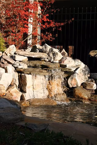 A small decorative rock waterfall flowing into a pond in a landscaped outdoor area with a red-leafed tree and a black fence behind.