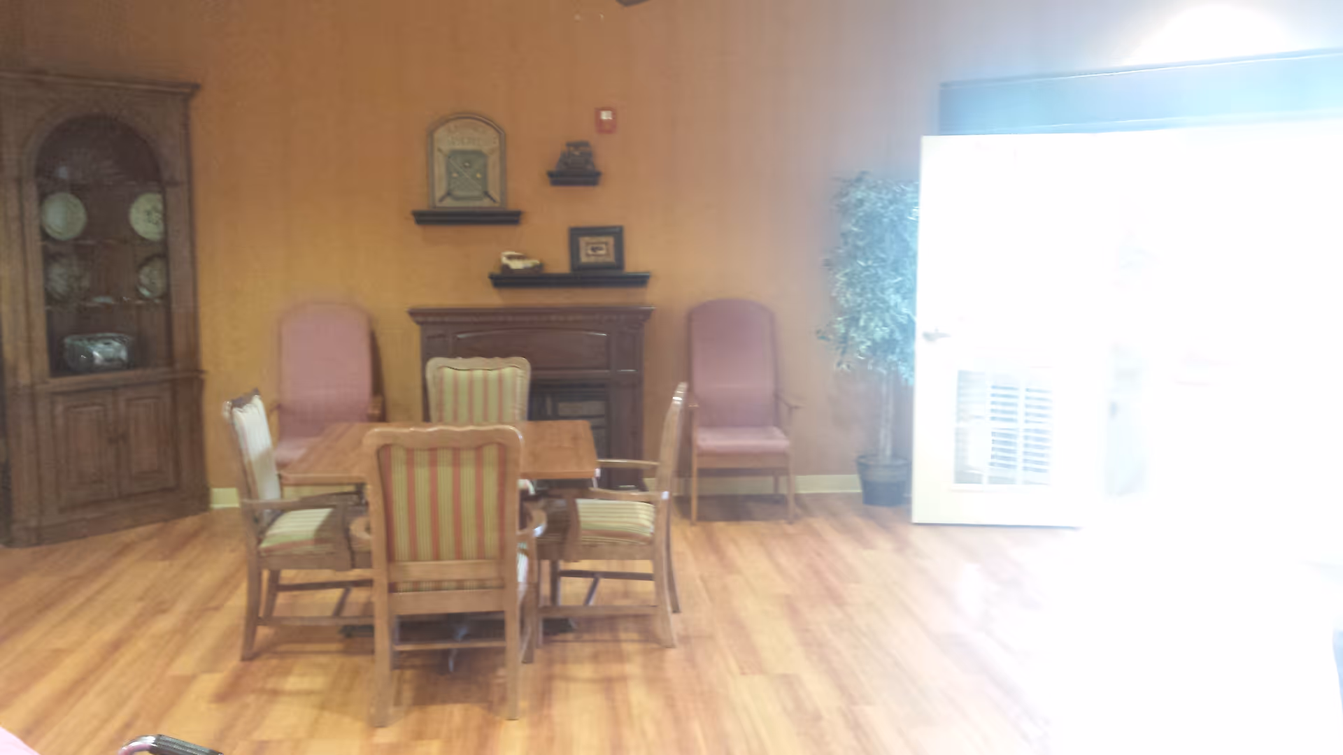 Small communal room with a wooden table surrounded by striped chairs, a cabinet and extra seating against a wood-paneled wall, and a bright glass door on the right.