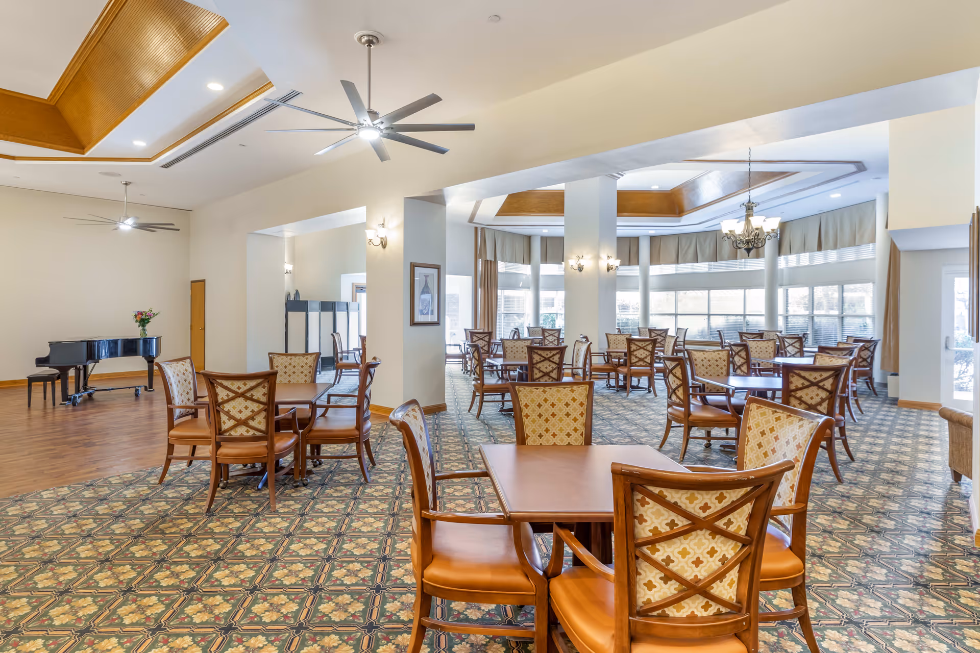 A spacious dining room with multiple wooden tables and cushioned chairs arranged neatly. The room features patterned carpet flooring, large windows with curtains allowing natural light, ceiling fans, and a chandelier. A black grand piano is visible in the corner on a wooden floor section.