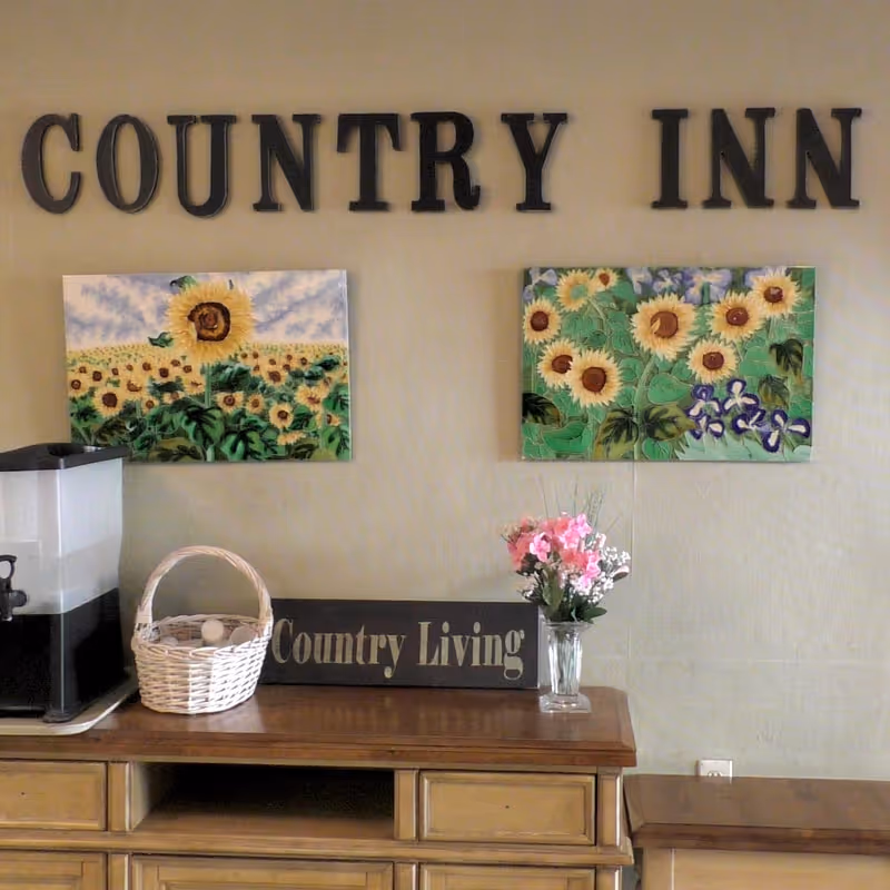 Interior wall reading 'COUNTRY INN' above sunflower paintings and a table with a coffee dispenser, wicker basket, 'Country Living' sign, and a vase of flowers.