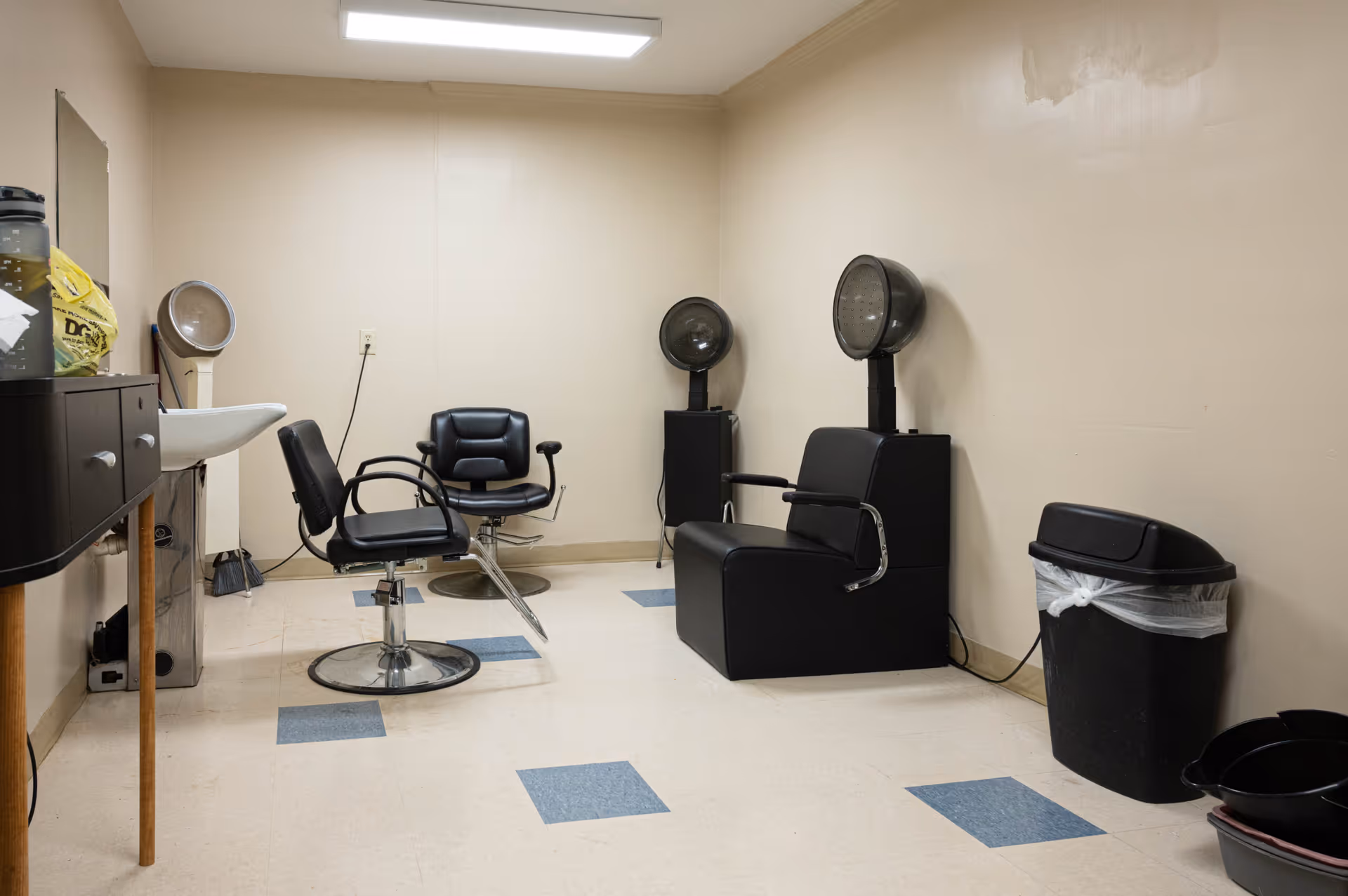 Small salon room with styling chairs, hooded hair dryers, a sink, and a trash can.