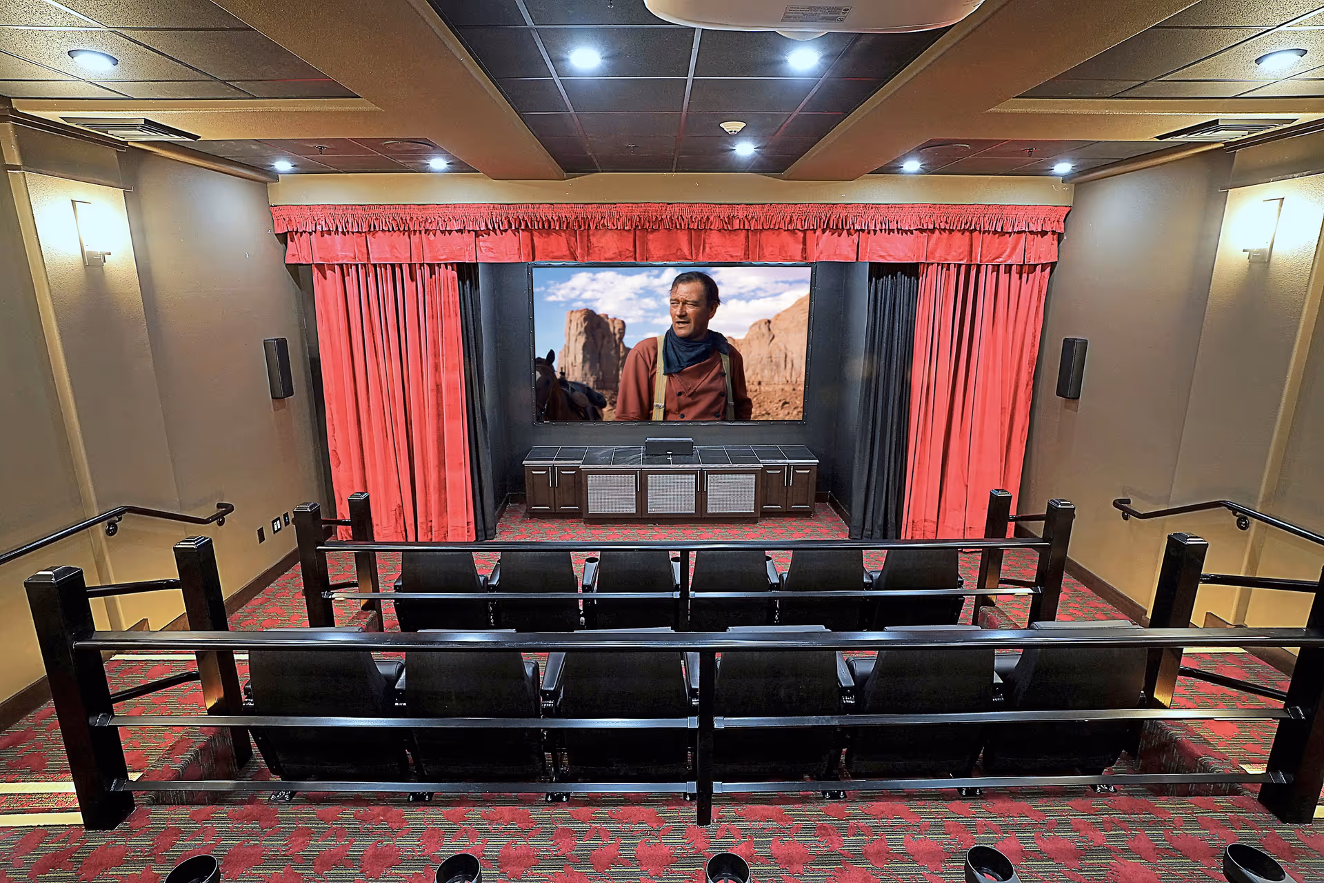 Small private theater room with tiered rows of seats facing a screen framed by red curtains showing a man in a desert scene.