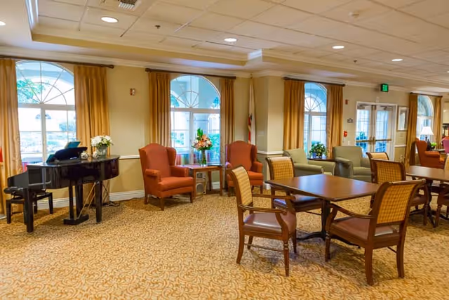 A bright and spacious common area in a senior living facility featuring large arched windows with yellow curtains, several comfortable armchairs, wooden tables with chairs, a black grand piano, and floral arrangements on tables. The room has a patterned carpet and recessed ceiling lights.