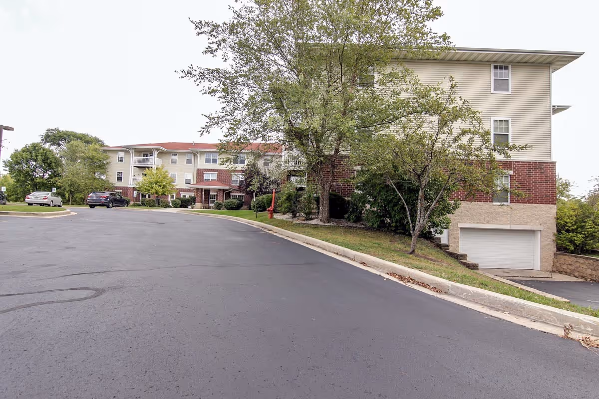 Exterior view of Bell Tower Place Senior Apartments showing a three-story building with beige siding and red brick accents. There are trees and shrubs around the building, a paved driveway, and a few parked cars visible.