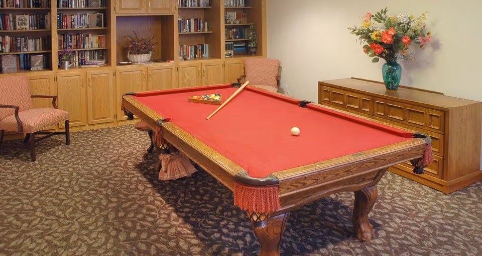 A carpeted common room featuring a red-felt pool table, bookshelves, upholstered chairs, and a sideboard with a vase of flowers.