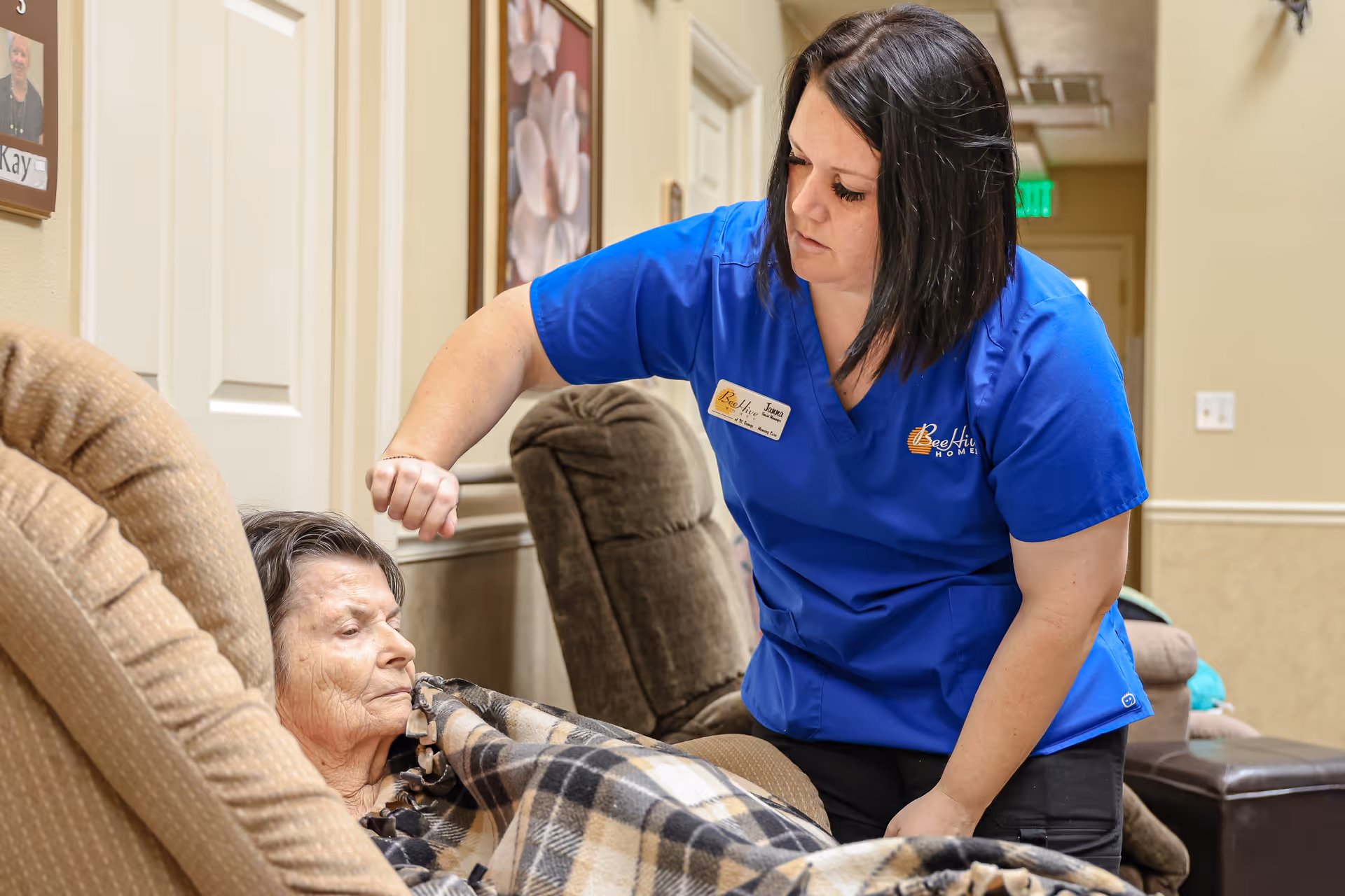 A caregiver in a blue uniform with a Beehive Homes logo is gently interacting with an elderly woman who is resting in a recliner chair covered with a plaid blanket in a cozy indoor setting.