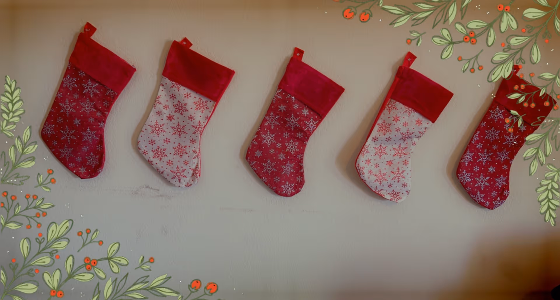 Five red and white Christmas stockings with snowflake patterns hanging on a wall, decorated with illustrated green leaves and red berries around the edges.