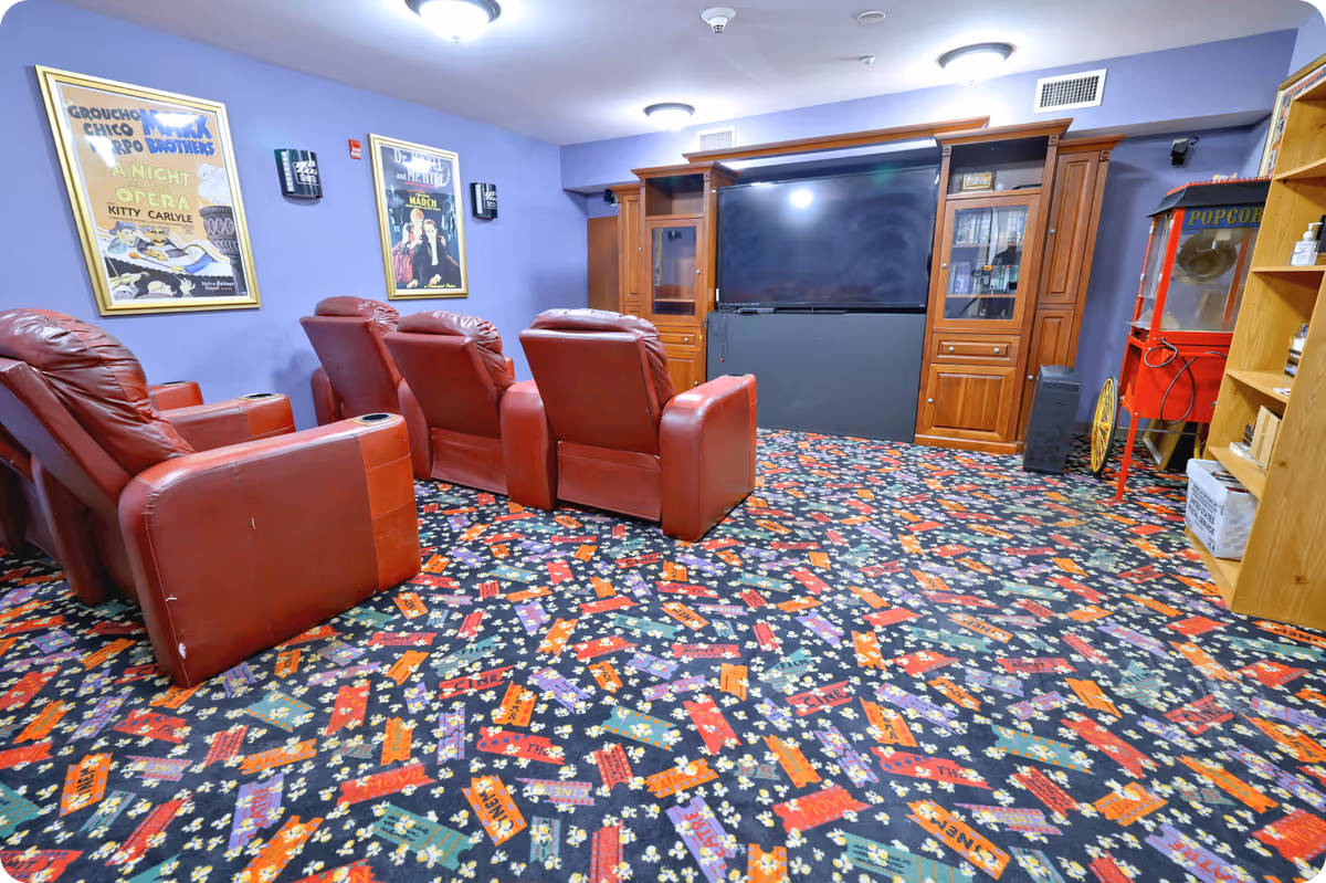 A cozy home theater room with four red leather recliner chairs arranged in two rows facing a large flat-screen TV mounted on a wooden entertainment center. The room has purple walls decorated with framed vintage movie posters and a colorful carpet with popcorn and movie ticket designs. A red popcorn machine and a wooden shelf with various items are visible on the right side.