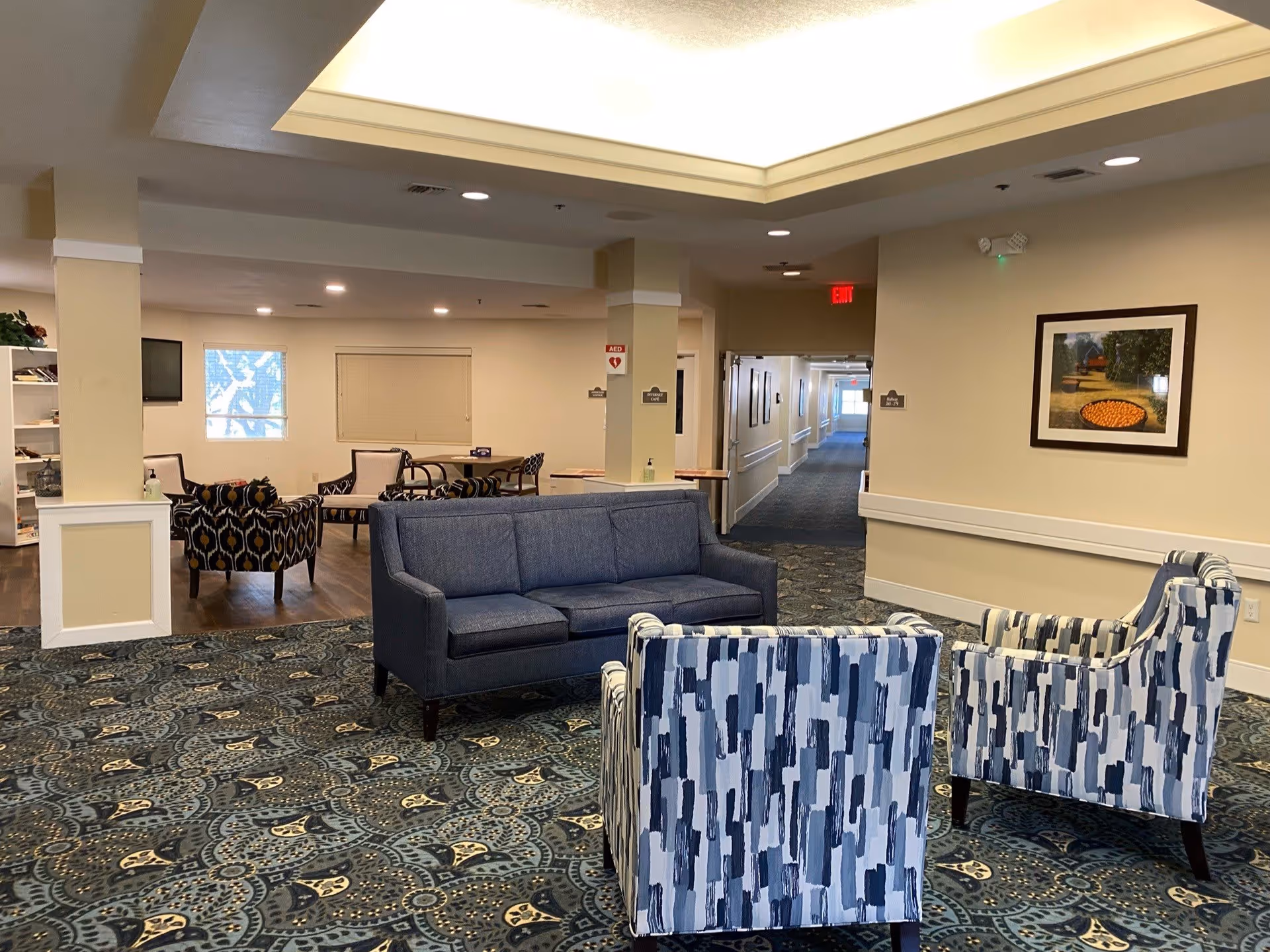 A senior living facility common area with a blue sofa and two patterned armchairs on a decorative carpet. In the background, there are additional seating areas with chairs and tables, a hallway leading to other rooms, and a framed picture on the wall. The space is well-lit with ceiling lights and has beige walls.