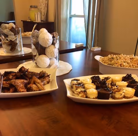 Plates of appetizers and small desserts on a wooden dining table with a decorative glass jar centerpiece and a window with curtains in the background.