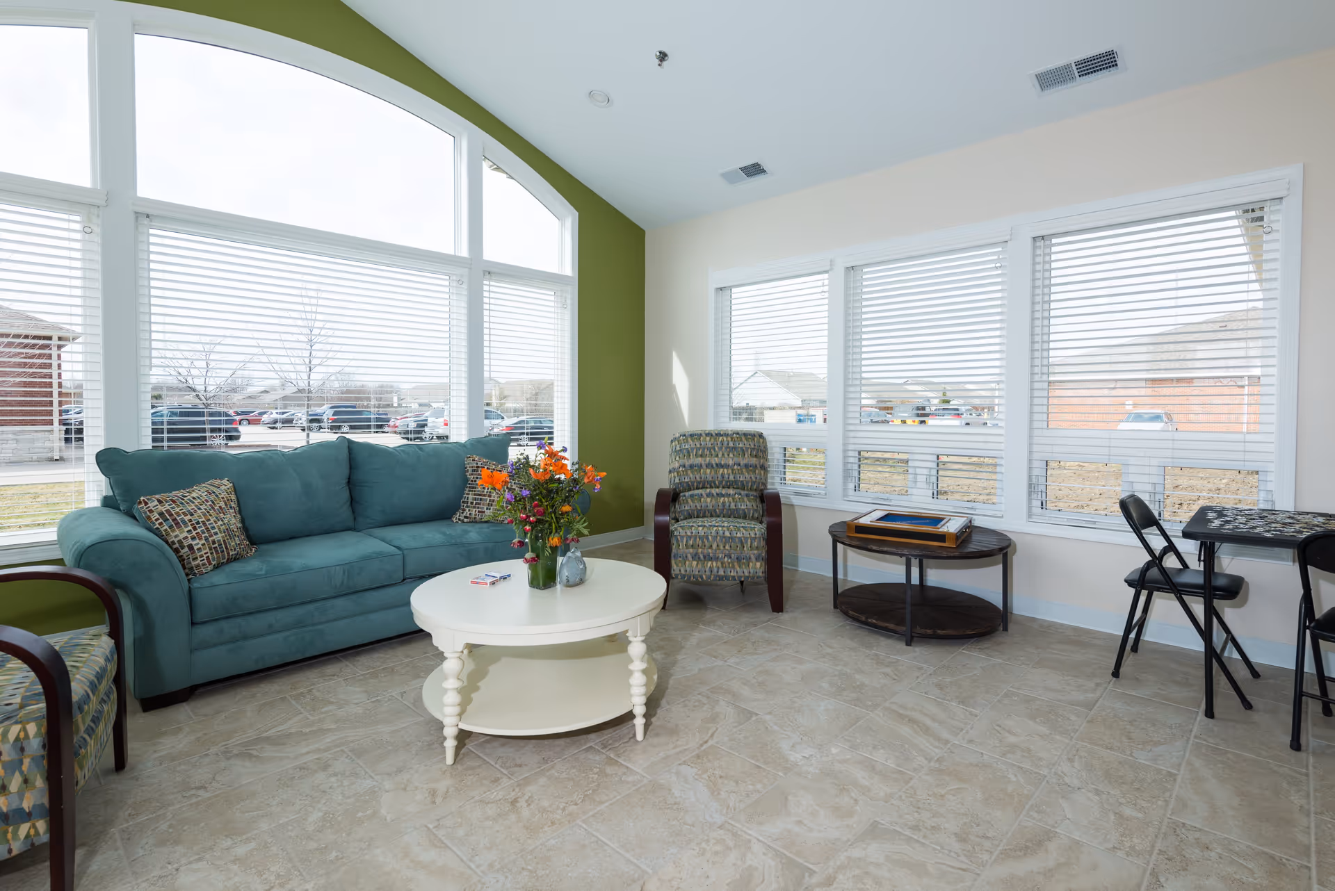 Bright sitting room with a teal sofa, patterned armchairs, a round coffee table with flowers, and large windows letting in daylight.