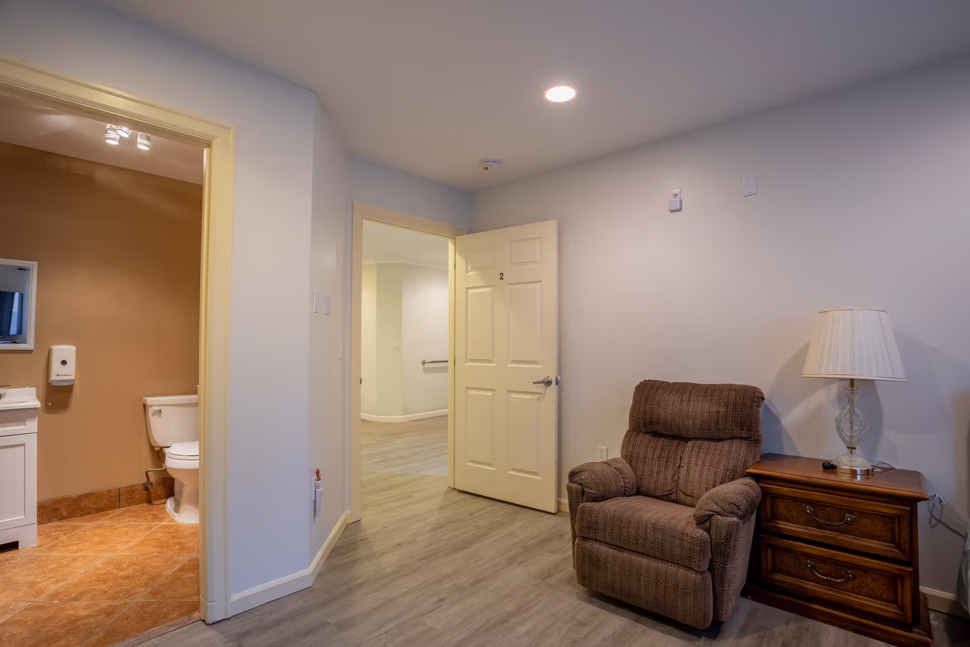 Interior view of a room in a care home showing a brown recliner chair next to a wooden side table with a lamp. The room has light-colored walls and wood flooring. An open door labeled with the number 2 leads to a hallway, and another open door reveals a bathroom with a toilet and sink.