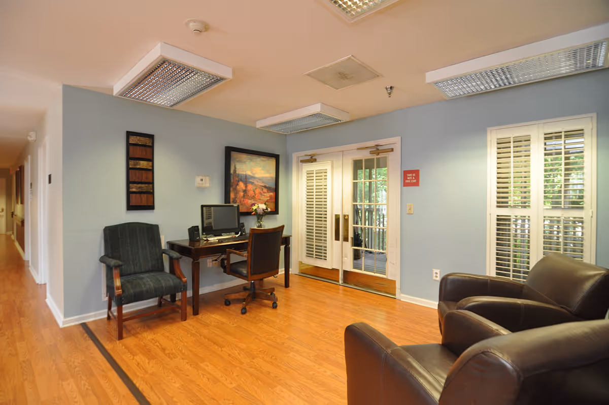 A cozy interior room with light blue walls and wooden flooring. The room features a small desk with a computer and a chair, a green upholstered armchair, and two dark brown leather armchairs. There are two white double doors with glass panels and white shutters on the windows. The ceiling has fluorescent lights, and there is a framed painting and decorative wall art on the walls.