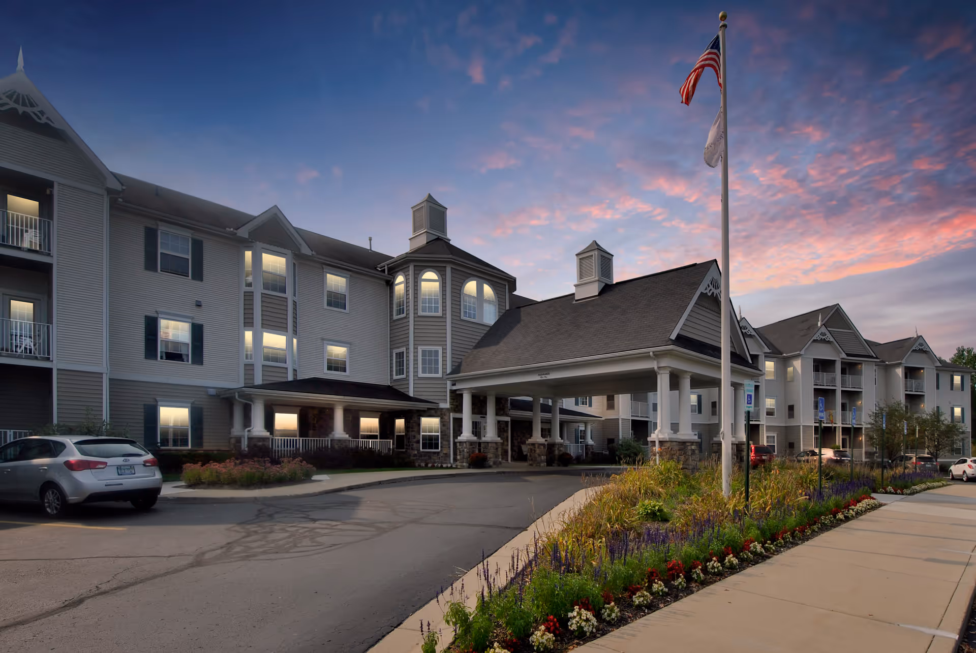 Exterior view of a multi-story senior living facility named Oakhaven Manor during sunset, featuring a covered entrance with columns, an American flag on a flagpole, landscaped flower beds, parked cars, and a partly cloudy sky with pink and purple hues.