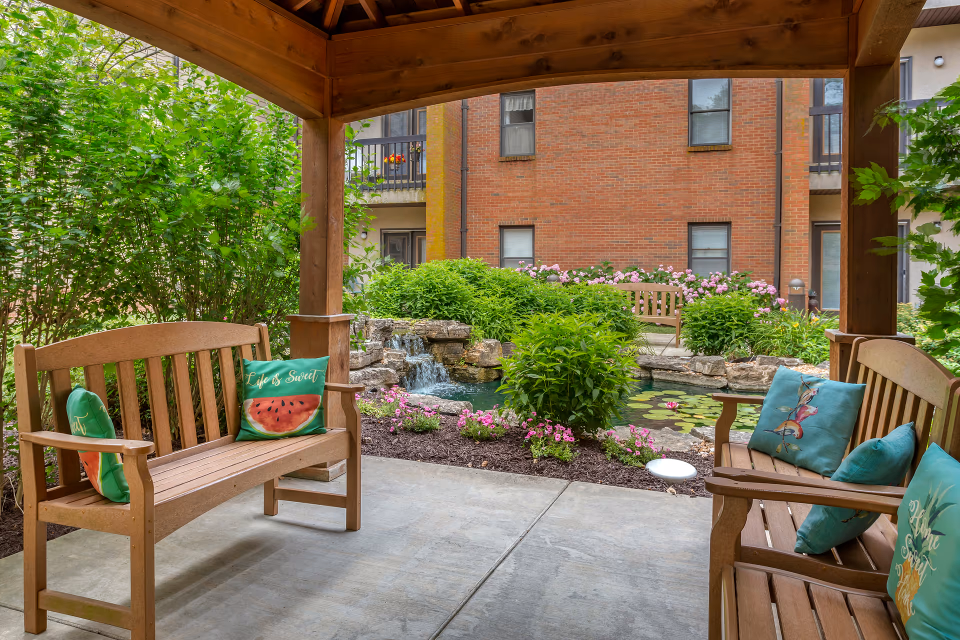 Covered courtyard seating area with wooden benches and colorful pillows overlooking a landscaped pond and small waterfall with a brick building in the background.