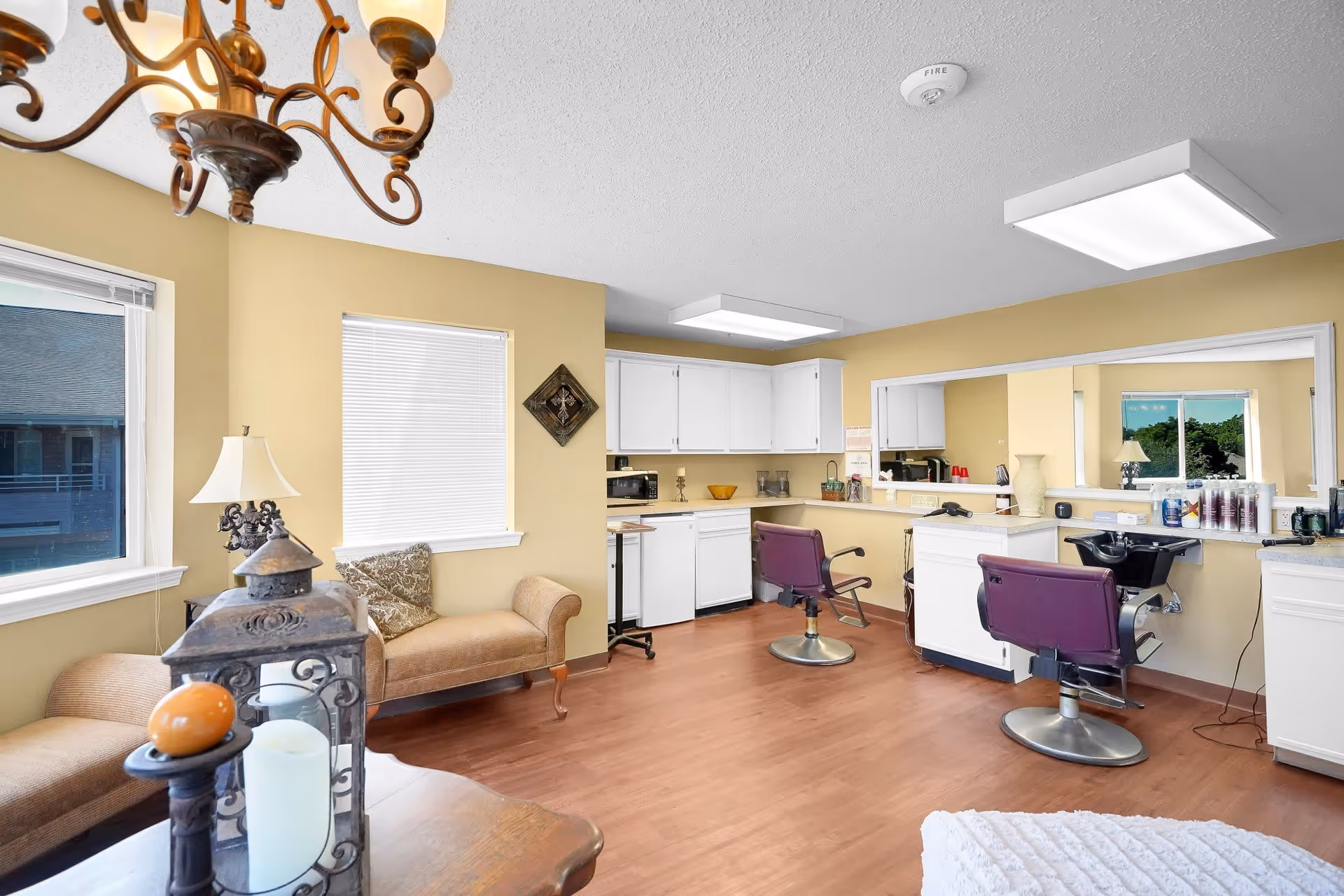 Interior view of a salon area in a senior living facility with two purple salon chairs in front of a large mirror, white cabinetry, and a washing station. The room has beige walls, wood flooring, a chandelier, and a seating area with a cushioned bench and a lamp near two windows.