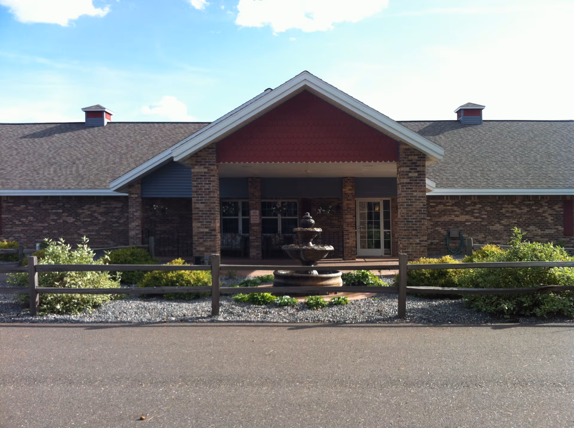 Front exterior view of a single-story brick building with a peaked roof and a covered entrance. There is a three-tiered water fountain in front of the entrance, surrounded by greenery and a wooden fence. The sky is partly cloudy.