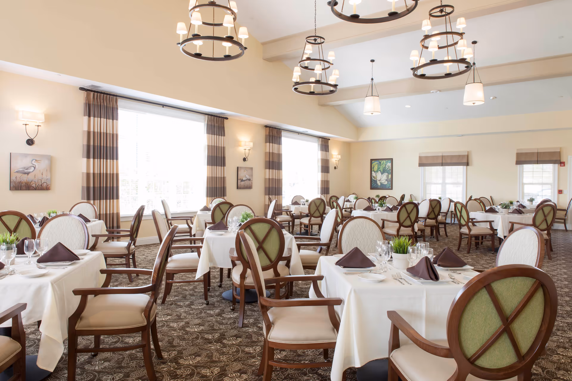 A bright and spacious dining room with multiple tables covered in white tablecloths, each set with glassware, plates, silverware, and brown folded napkins. The chairs have wooden frames with green and beige upholstered backs and seats. Large windows with striped curtains allow natural light to fill the room. The ceiling features several elegant chandeliers and pendant lights. Artwork decorates the walls, and the carpet has a detailed pattern.