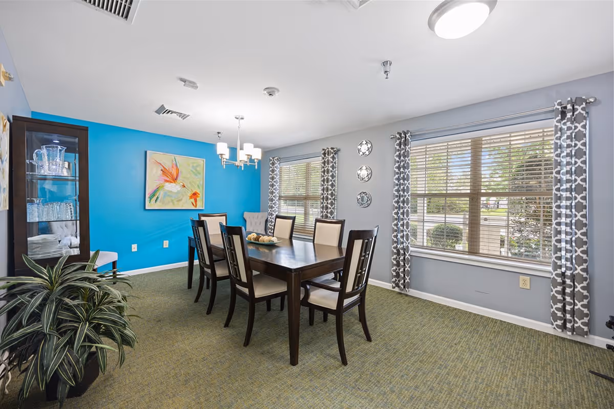 Bright communal dining room with a dark wood table and chairs, blue accent wall, large windows with patterned curtains, and a china cabinet.