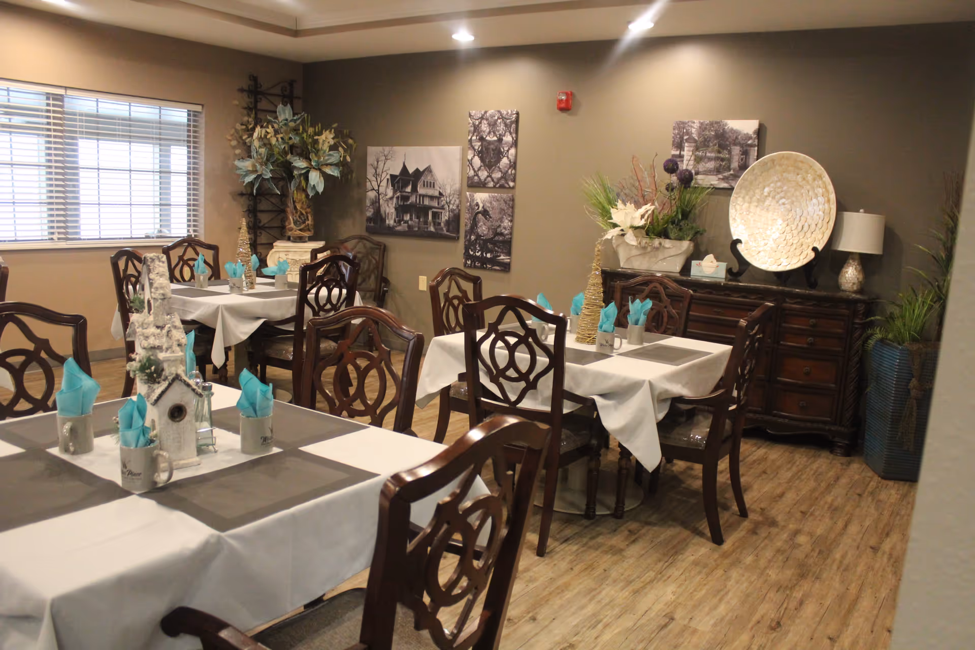 Interior view of a dining room at Weller Place Retirement Center featuring multiple tables covered with white tablecloths and gray placemats, each set with turquoise napkins in mugs. The room has wooden chairs with decorative backs, wood flooring, and beige walls adorned with black and white framed pictures. A wooden sideboard with decorative plants and a large ornamental plate is visible against one wall.