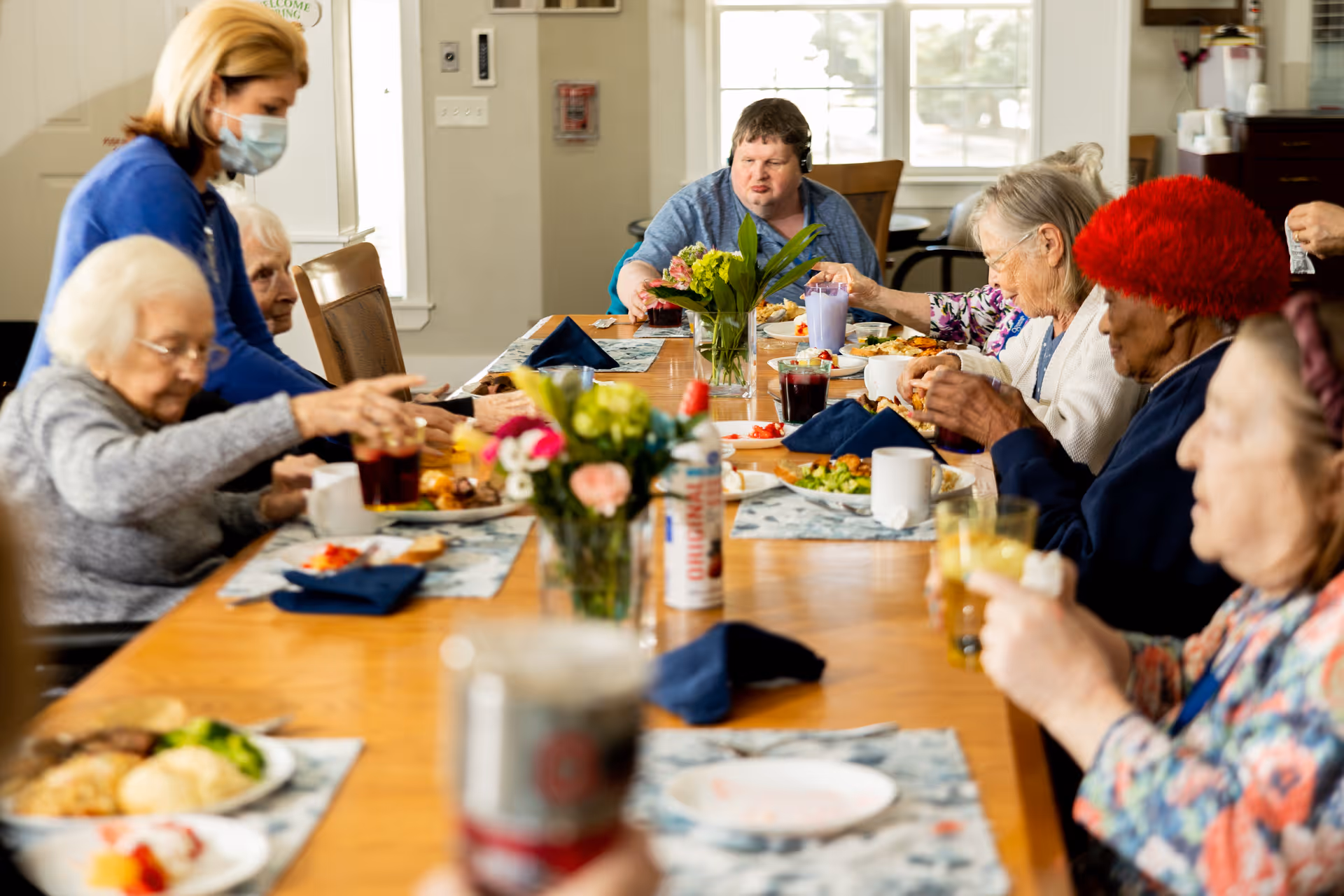 A group of older adults and a masked caregiver seated around a long dining table sharing a meal.
