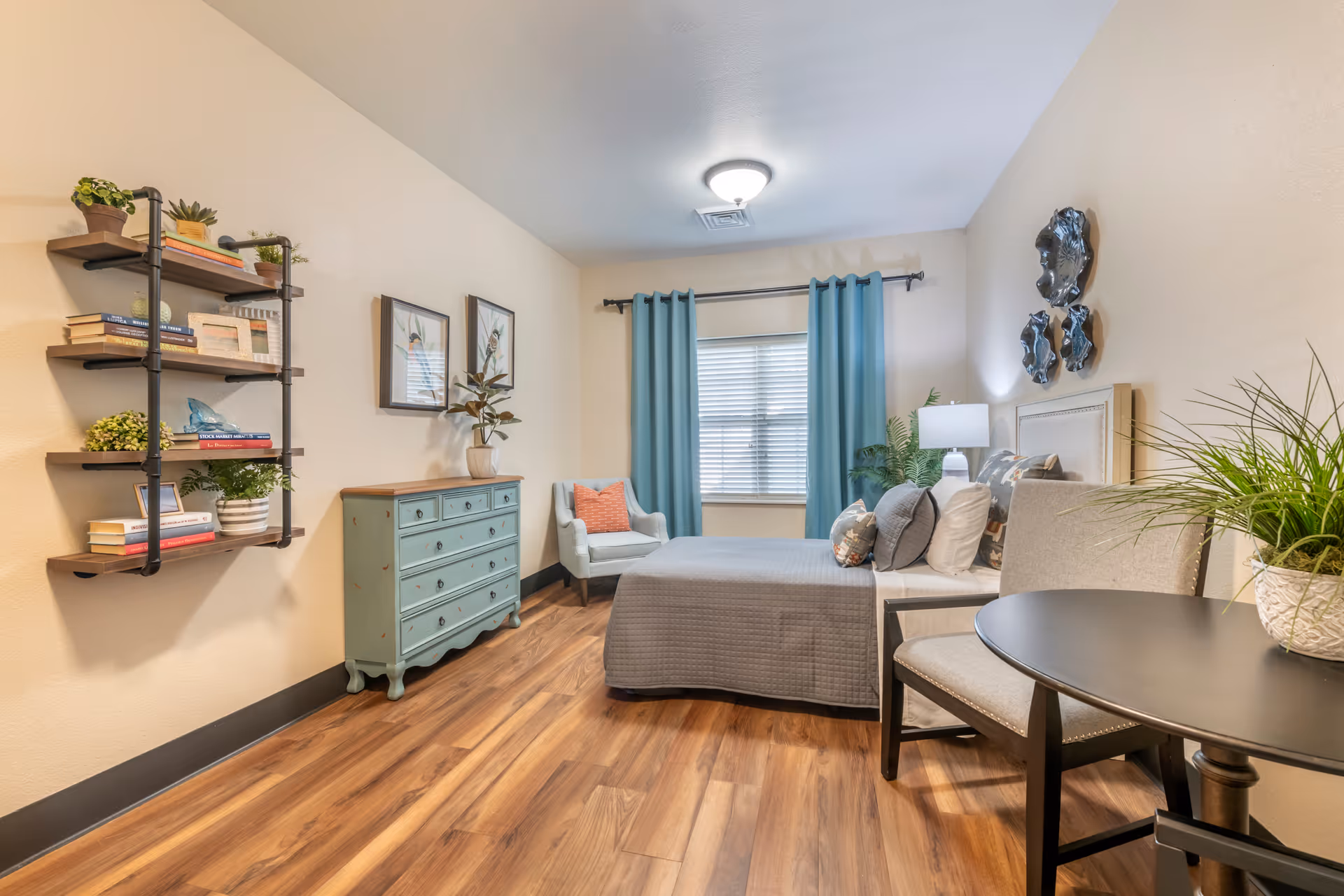 A cozy bedroom with a bed covered in gray bedding and multiple pillows, a light blue armchair with an orange pillow, a teal dresser, wall-mounted shelves with books and plants, a round black table with a beige chair, and blue curtains on the window. The room has wooden flooring and neutral-colored walls with decorative wall art.