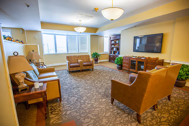 Bright communal living room with sofas and armchairs arranged on a patterned rug, a wall-mounted TV, bookshelf and large windows.