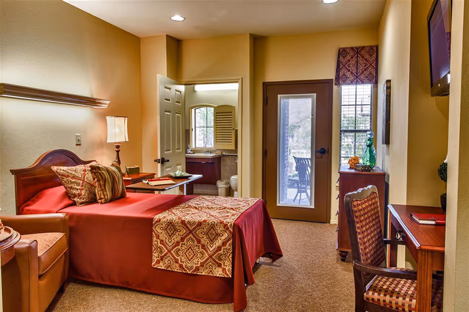 Warmly lit bedroom with a single bed, armchair, desk, and a door to a small patio with a bathroom visible through an open door.