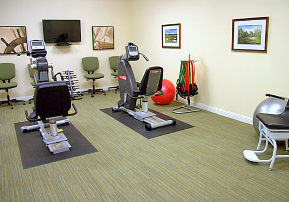 Small fitness room with two recumbent exercise bikes, free weights, exercise ball, resistance bands, chairs and a wall-mounted TV.