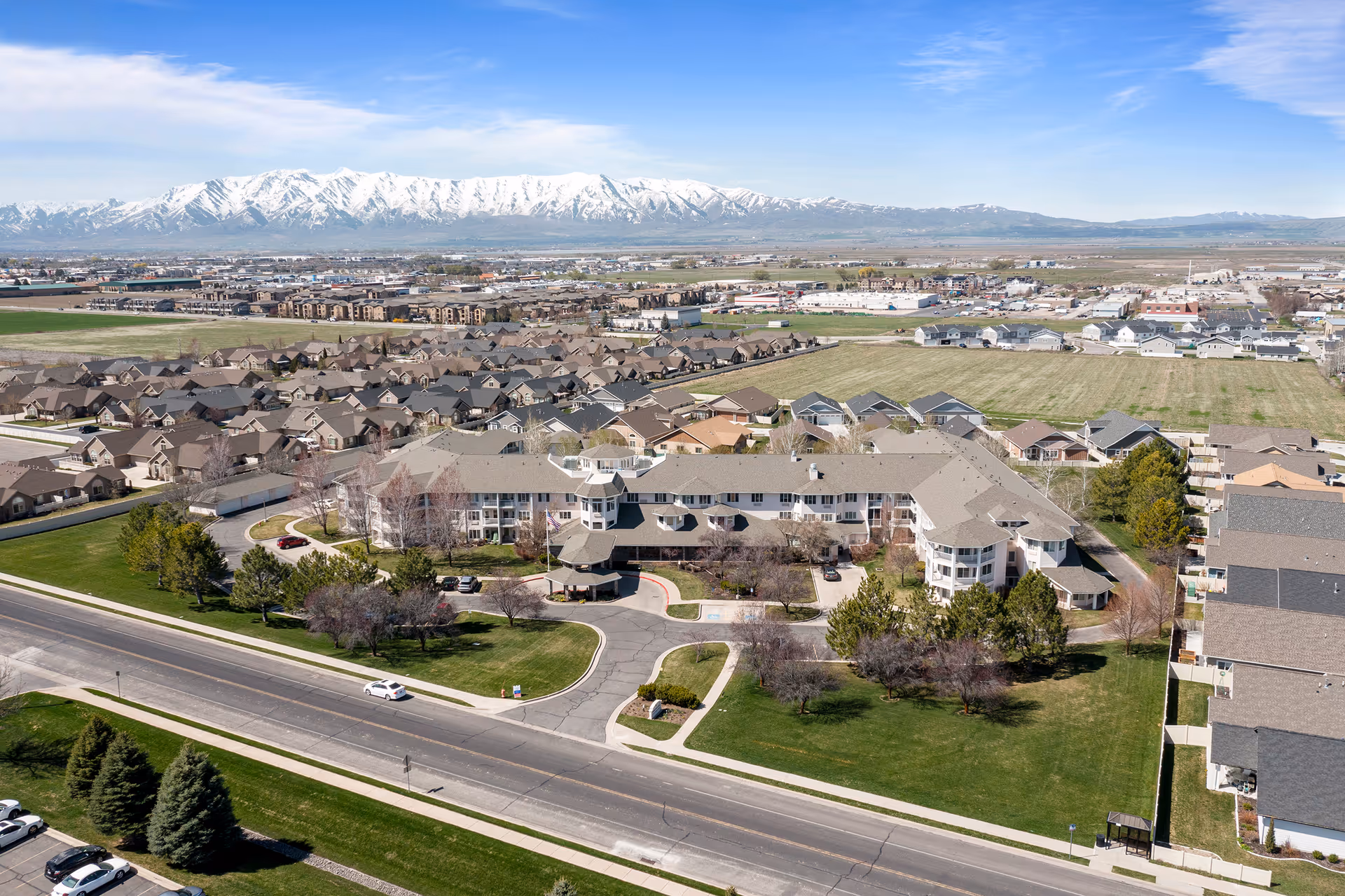 Aerial view of Pioneer Valley Lodge Gracious Retirement Living facility surrounded by residential houses and green lawns, with snow-capped mountains in the background under a clear blue sky.