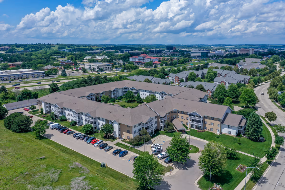 Aerial view of a large senior housing apartment community with multiple connected buildings, surrounded by green lawns, trees, and parking lots. The sky is partly cloudy with blue patches.
